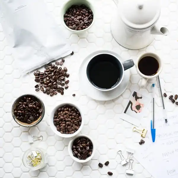 Cups of black coffee, small bowls of beans and a white coffee bag on a white tiled table