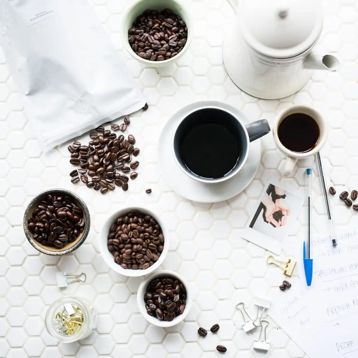 Cups of black coffee, small bowls of beans and a white coffee bag on a white tiled table