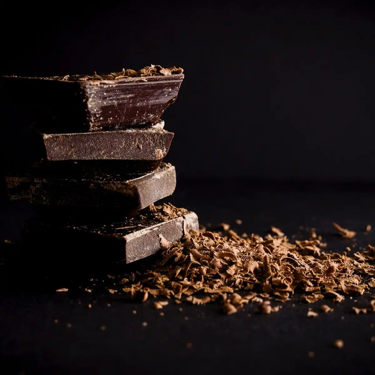 A stack of broken dark chocolate pieces with shavings on a black surface