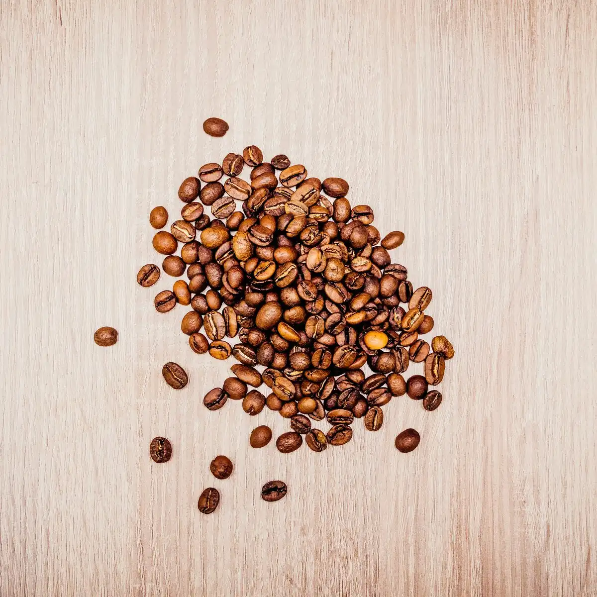 A loose pile of medium-brown roasted coffee beans on a pale wooden board