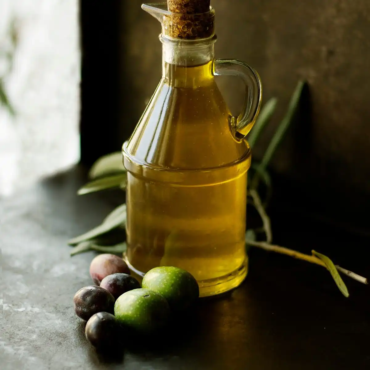 A glass cruet of olive oil with a cork stopper next to green and black olives