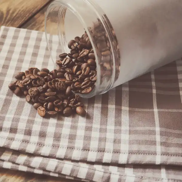 Roasted coffee beans spilling from a tipped glass jar onto a checked cloth