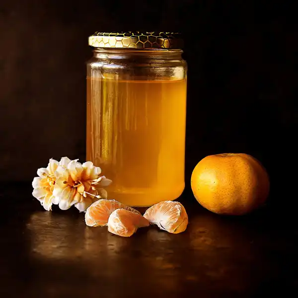 A glass jar of golden honey beside a peeled mandarin on a dark table