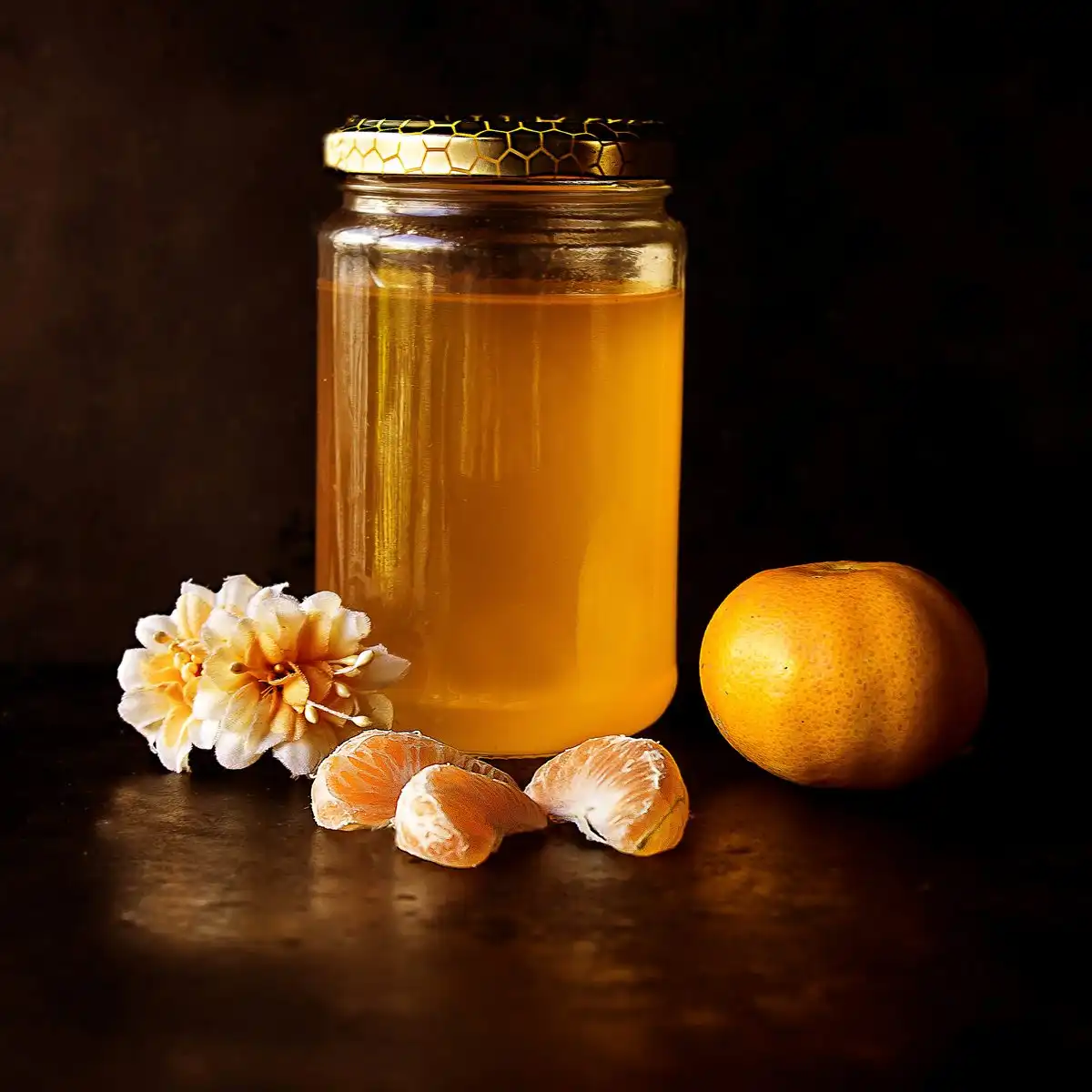 A glass jar of golden honey beside a peeled mandarin on a dark table